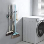Soporte organizador de acero inoxidable para escobas y utensilios colgado en pared gris, junto a lavadora blanca.