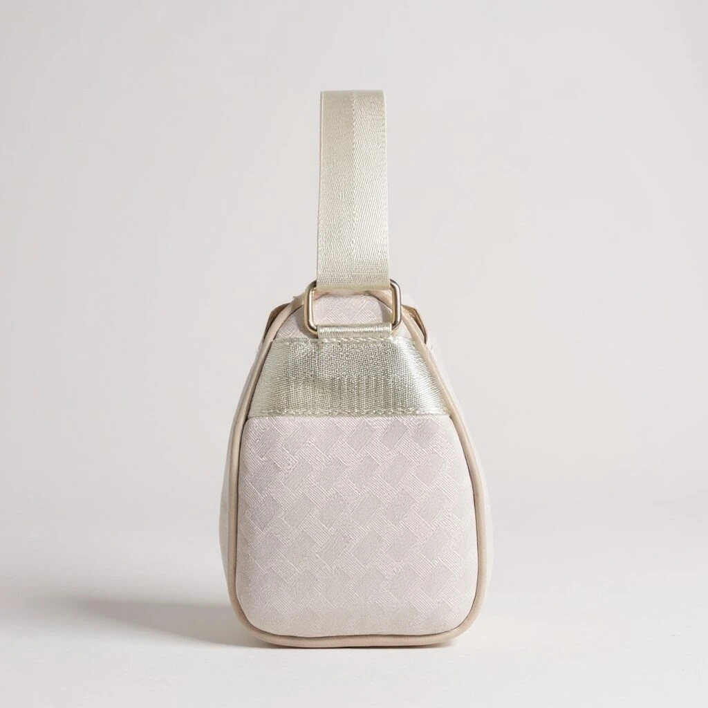Vista lateral de bolso dama Dana beige con textura tejida sutil, ribetes y correa de mano en tono dorado claro, herrajes metálicos.
