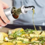Botella de vidrio con dispensador metálico rociando aceite sobre papas y hierbas frescas.