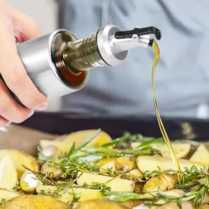 Botella de vidrio con dispensador metálico rociando aceite sobre papas y hierbas frescas.