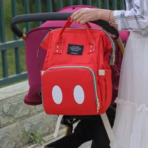Morral pañalera roja estilo Mickey Mouse colgado en manija de coche de bebé, mujer de vestido blanco lo sujeta.