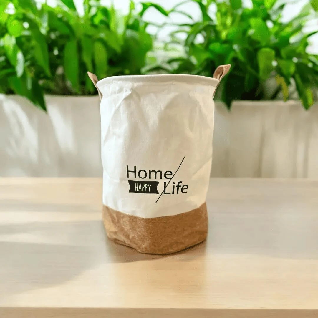 Cesto organizador de tela lona blanco y base marrón, con asas y texto "Home Happy Life", sobre una mesa con plantas al fondo.