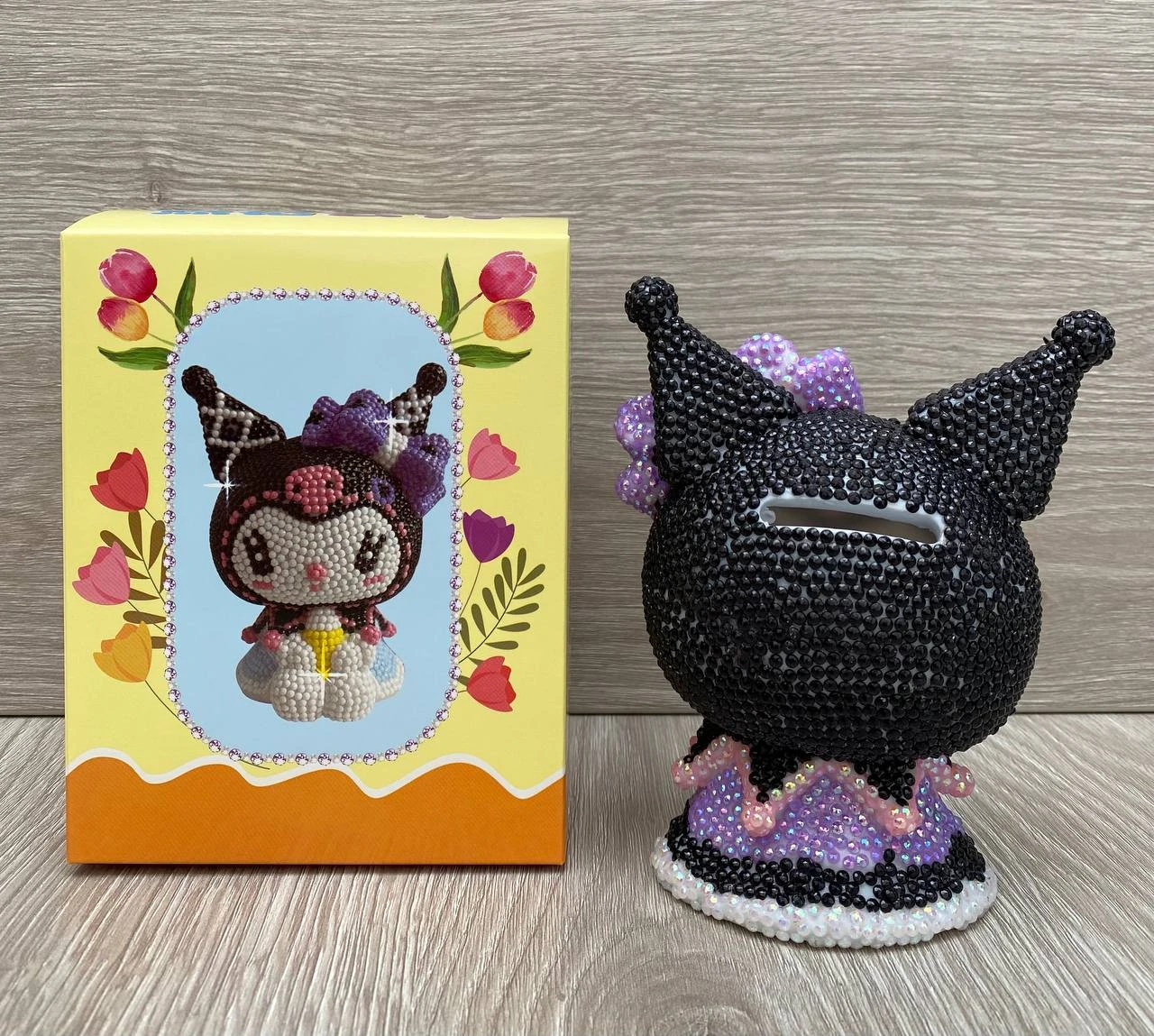 Alcancía Kuromi negra con pedrería y caja amarilla decorada con flores, mostrando el kit de pintura de diamantes.