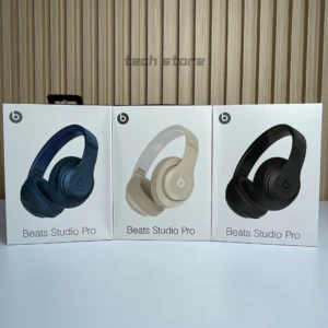 Tres cajas de Beats Studio Pro: azul oscuro, beige claro y negro, sobre una superficie blanca y fondo de listones de madera.