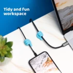 Dos sujetacables de silicona azul con forma de mano pulgar arriba sujetan cables de carga de celular y portátil en escritorio blanco.