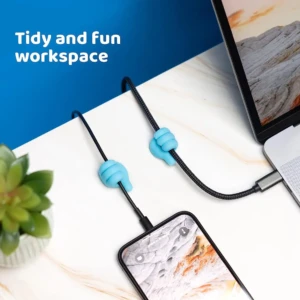 Dos sujetacables de silicona azul con forma de mano pulgar arriba sujetan cables de carga de celular y portátil en escritorio blanco.