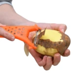 Pelador naranja con perforaciones usando su cuchilla para quitarle la cáscara a una papa cruda.