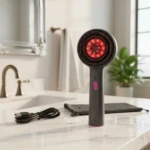 Cepillo masajeador capilar gris oscuro con luz LED roja circular, incluye cable de carga USB, estuche y protector sobre encimera de baño de mármol.