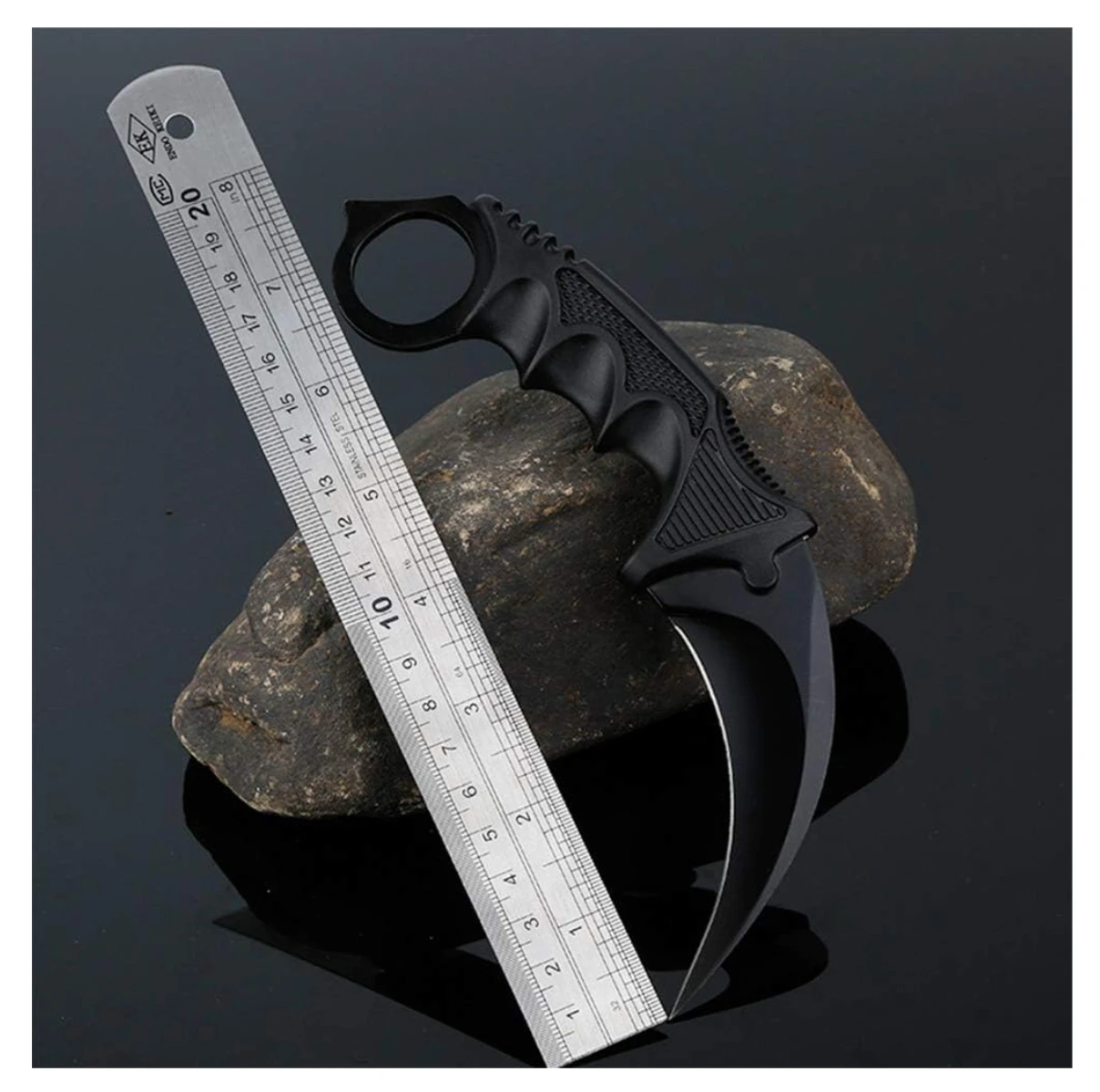 Cuchillo Karambit táctico negro con hoja curva, junto a una regla metálica sobre una roca y fondo oscuro.