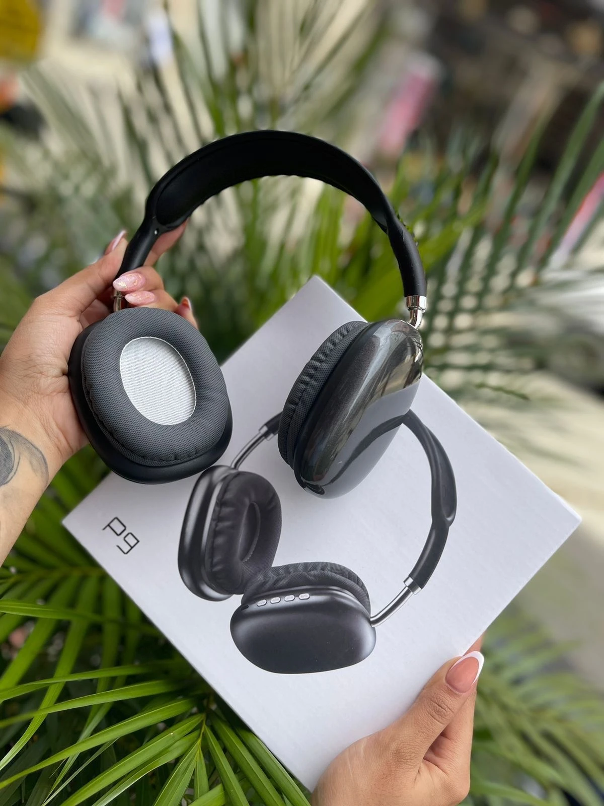 Auriculares inalámbricos Diadema P9 negros sostenidos sobre su caja blanca, con fondo de plantas verdes.