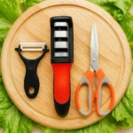 Afilador rojo y negro, tijeras naranjas, pelador negro sobre tabla de madera y hojas de lechuga.