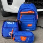Set escolar Totto azul vibrante: morral, lonchera y estuche. Detalles en naranja neón y cremalleras visibles. Fondo exterior blanco de vehículo.