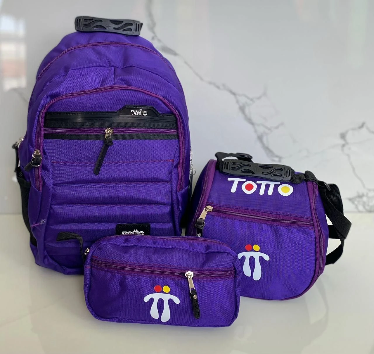 Set escolar Totto morado: morral grande, lonchera y estuche pequeño con logo blanco y detalles en rojo y amarillo.