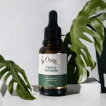 Serum Origenes para cejas y pestañas, frasco de vidrio ámbar con gotero, hojas verdes de monstera.