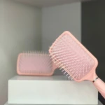 Cepillo plano para cabello rosa pálido con cerdas blancas, un cepillo enfocado y otro desenfocado al fondo sobre caja blanca.
