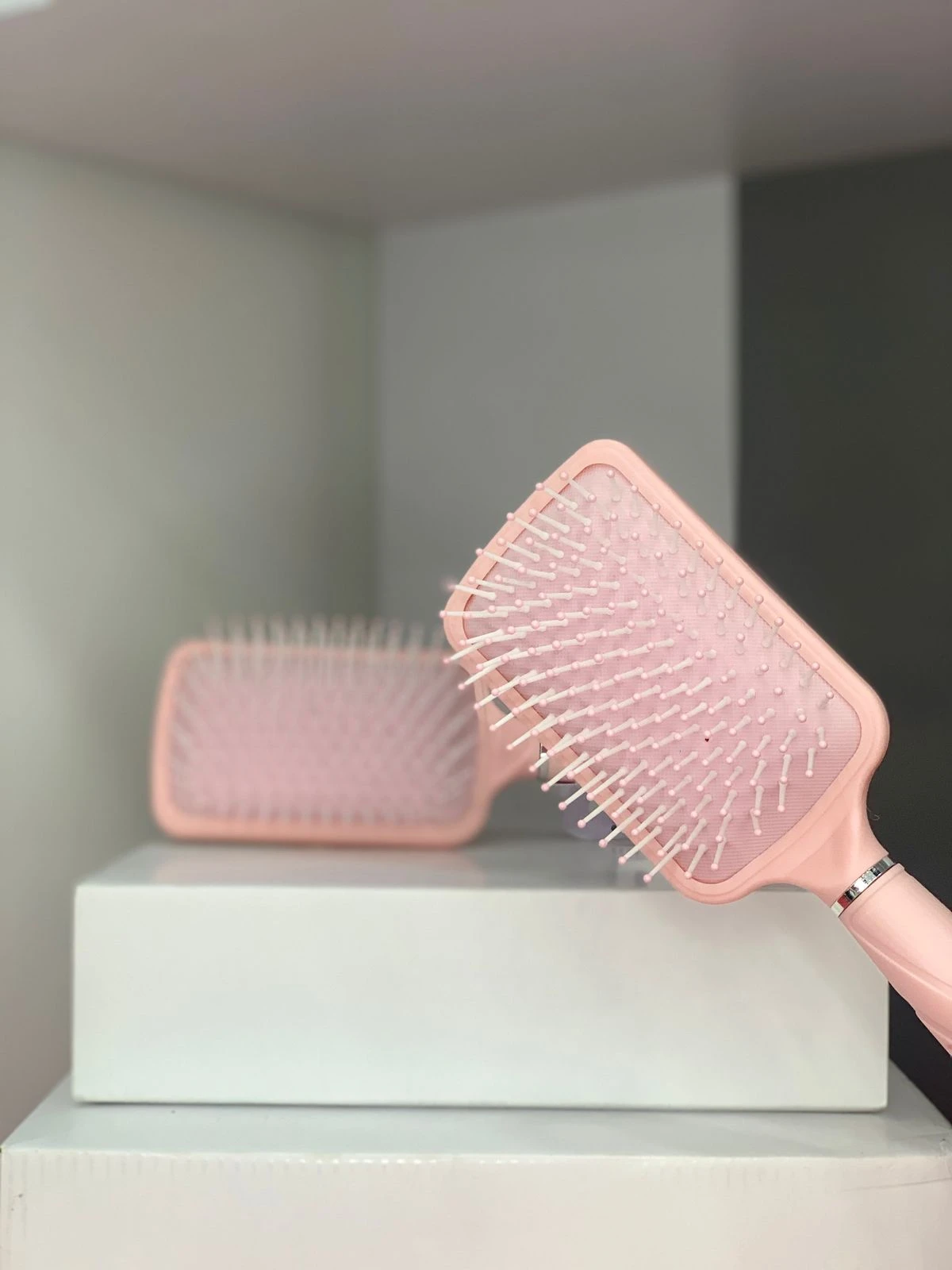 Cepillo plano para cabello rosa pálido con cerdas blancas, un cepillo enfocado y otro desenfocado al fondo sobre caja blanca.