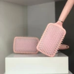Cepillo para cabello rosa claro modelo paleta con cerdas blancas, uno inclinado sobre cajas blancas con otro y parte de un tercero de fondo.