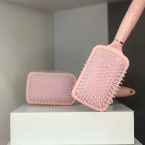 Cepillo para cabello rosa claro modelo paleta con cerdas blancas, uno inclinado sobre cajas blancas con otro y parte de un tercero de fondo.