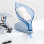 Jabonera azul con forma de hoja y base gris, con jabón blanco encima, sobre lavabo junto a grifería cromada.