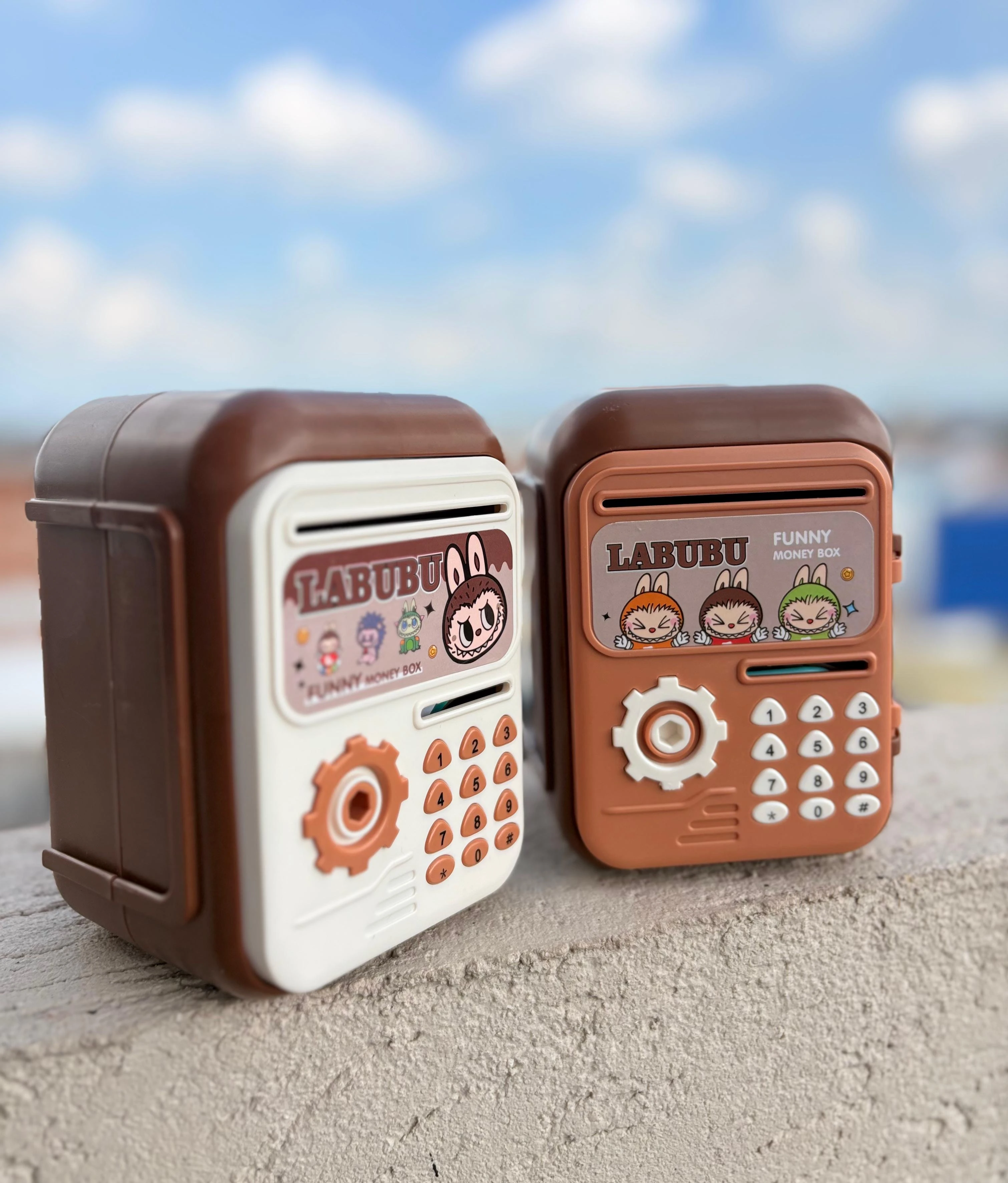 Set de dos alcancías electrónicas Labubu con teclado numérico, una blanca y otra terracota, sobre superficie rugosa con cielo azul de fondo.