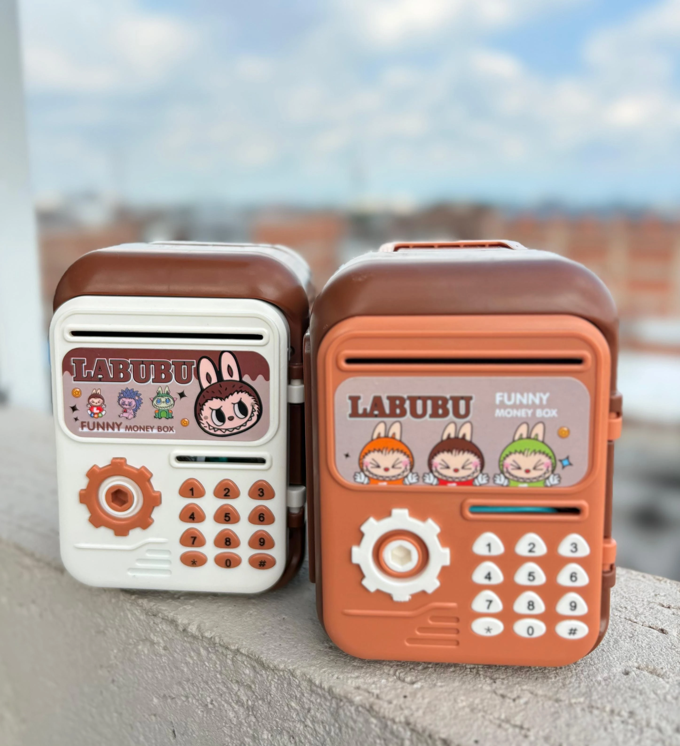 Dos alcancías electrónicas LABUBU estilo caja fuerte, una blanca y otra naranja, sobre superficie rugosa con cielo de fondo.