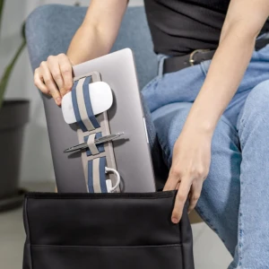 Organizador adhesivo con bandas elásticas azul y beige sujetando ratón, bolígrafo y cable a la tapa de un computador portátil gris antes de guardarlo en bolso negro.