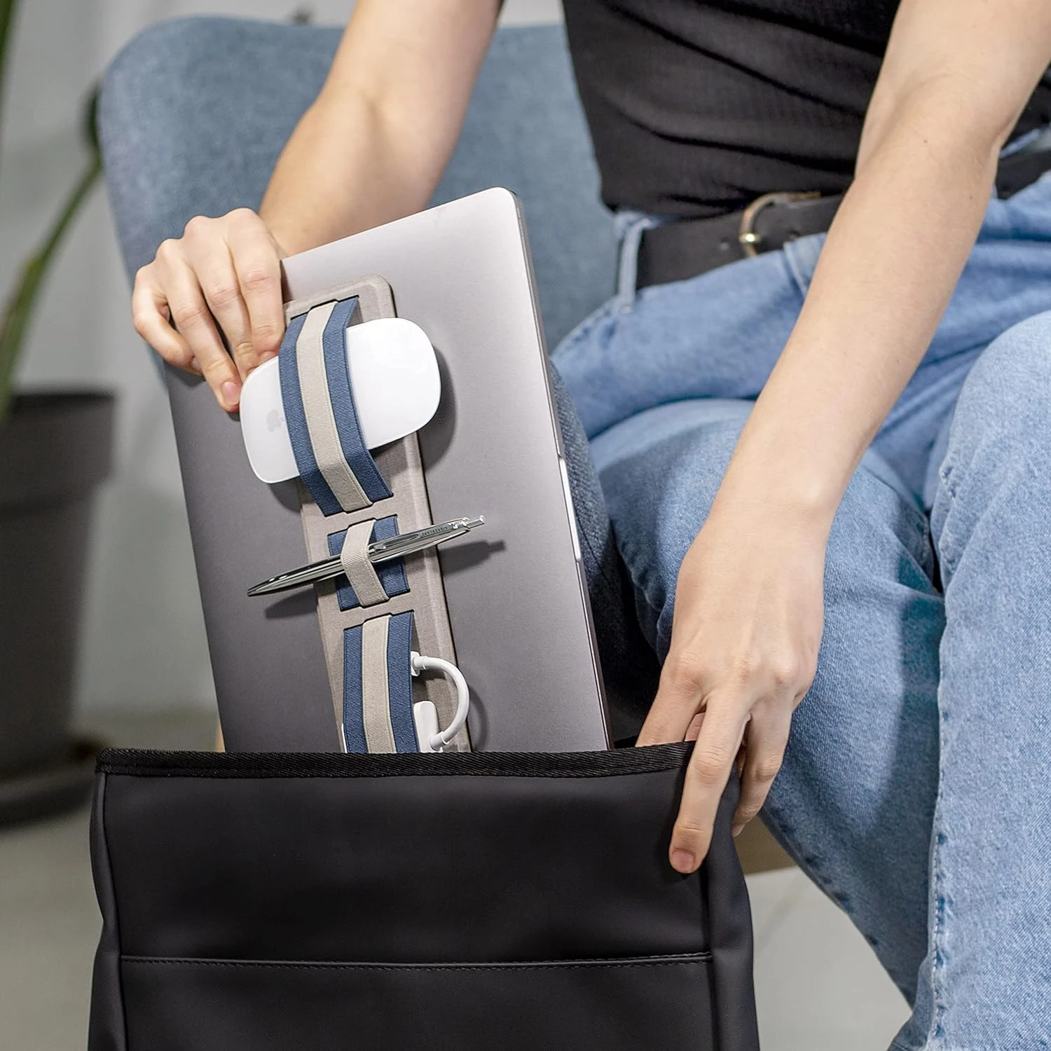 Organizador adhesivo con bandas elásticas azul y beige sujetando ratón, bolígrafo y cable a la tapa de un computador portátil gris antes de guardarlo en bolso negro.