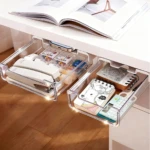 Dos cajones organizadores transparentes debajo de un escritorio blanco, llenos de suministros de oficina como grapadoras, notas y calculadora.