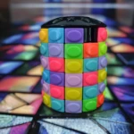 Cubo cilíndrico tipo rompecabezas con bloques sensoriales de colores neón y tapa negra, sobre fondo abstracto tipo vitral desenfocado.