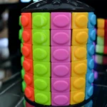 Cubo Rompecabezas cilíndrico de bloques apilables con caras en rojo, verde, rosa, amarillo y azul. Reflejo en base negra.