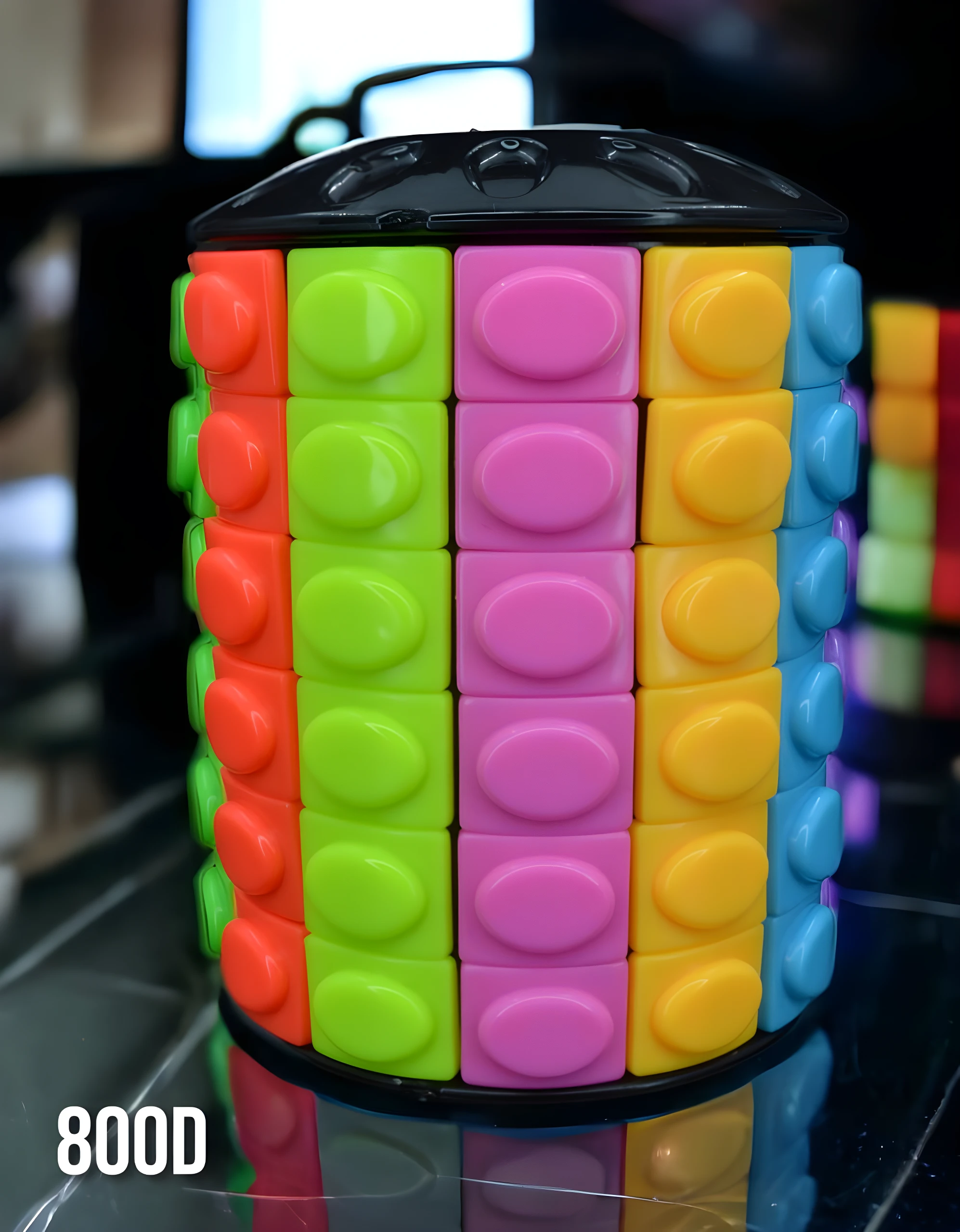 Cubo Rompecabezas cilíndrico de bloques apilables con caras en rojo, verde, rosa, amarillo y azul. Reflejo en base negra.
