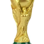 Alcancía dorada tipo trofeo Copa Mundial, con base verde y detalles del evento, ideal para coleccionistas.