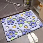 Alfombrilla de baño morada con diseño floral 3D, corazones amarillos y blancos, junto a pantuflas blancas en suelo de baldosa gris.