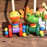 Juguete caminador de madera: oruga pato amarillo y oruga rana verde, con ruedas y anillos apilables de colores vivos sobre fondo de madera.