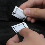 Manos separando dos parches adhesivos plateados texturizados sobre tela negra de malla, probablemente para reparar un armario organizador portátil.