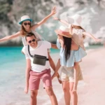 Hombre con camiseta blanca y bermudas rosas lleva riñonera impermeable negra cruzada en un día soleado en playa tropical.