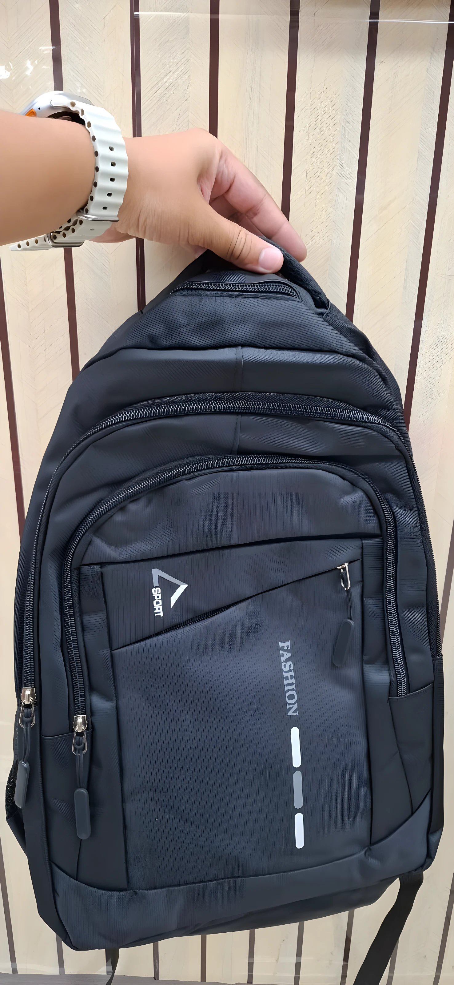 Mochila negra con logo "Fashion" y "Sport", ideal para escolar o viajero, sostenida por mano con reloj blanco.