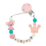 Sujetachupete de silicona rosa con clip, dije koala, corona, cuentas, y letras formando "BABY". Ideal para paseo y dentición.