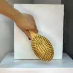 Mano sosteniendo cepillo de madera para cabello, con cerdas redondas, sobre fondo blanco y gris.