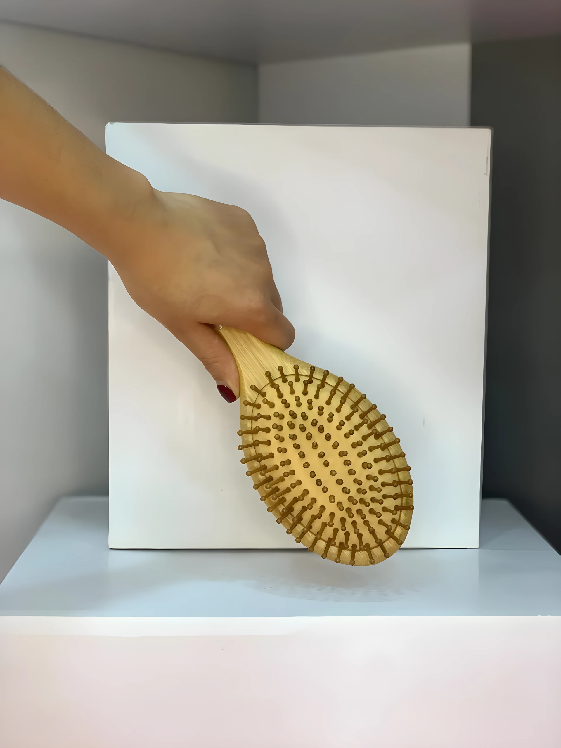 Mano sosteniendo cepillo de madera para cabello, con cerdas redondas, sobre fondo blanco y gris.