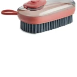 Cepillo limpiador con cerdas oscuras, cuerpo rojo terracota y tapa protectora transparente con botón superior. Incluye asa para colgar.