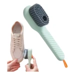 Cepillo limpiador de calzado verde menta con cerdas grises, dispensador de jabón integrado, usado sobre tenis blancos.