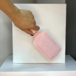 Mano sosteniendo un Cepillo Glam de paleta plano color rosa claro con cerdas blancas sobre fondo blanco.