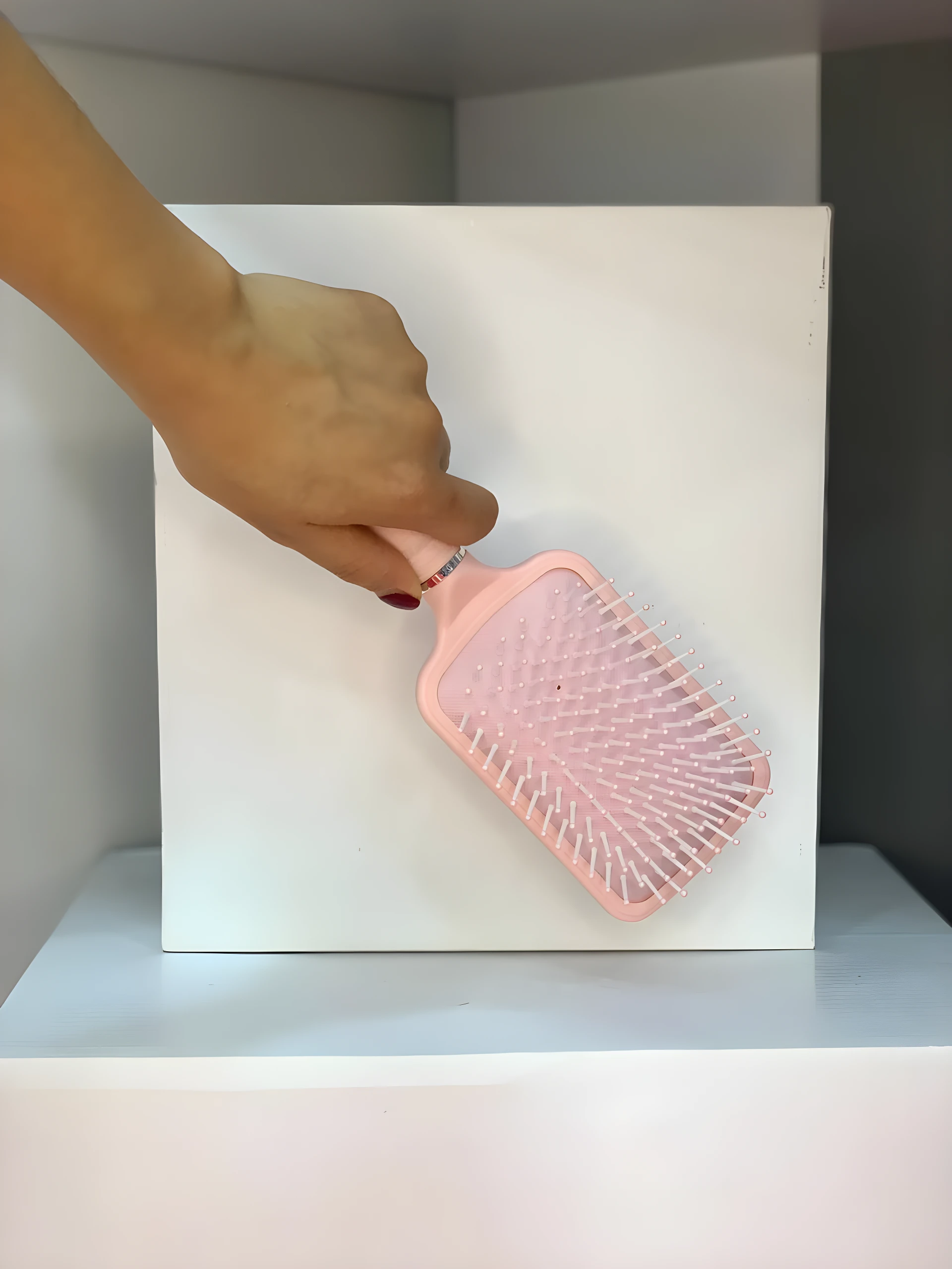 Mano sosteniendo un Cepillo Glam de paleta plano color rosa claro con cerdas blancas sobre fondo blanco.