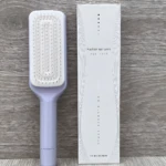 Cepillo peine retráctil morado de limpieza automática para todo tipo de cabello, en empaque blanco minimalista.