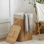 Cesta de ropa sucia de bambú con forro y tapa, con manta gris y silla de madera al lado.