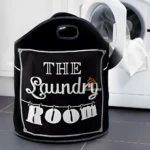 Cesta negra para ropa sucia con texto blanco "The Laundry Room", junto a lavadora.