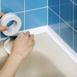 Aplicación de cinta selladora blanca impermeable de 32 mm en la esquina entre azulejo azul y superficie beige.