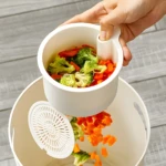 Colador blanco vertiendo brócoli y zanahoria picada en un tazón receptor, separando el líquido con un filtro.