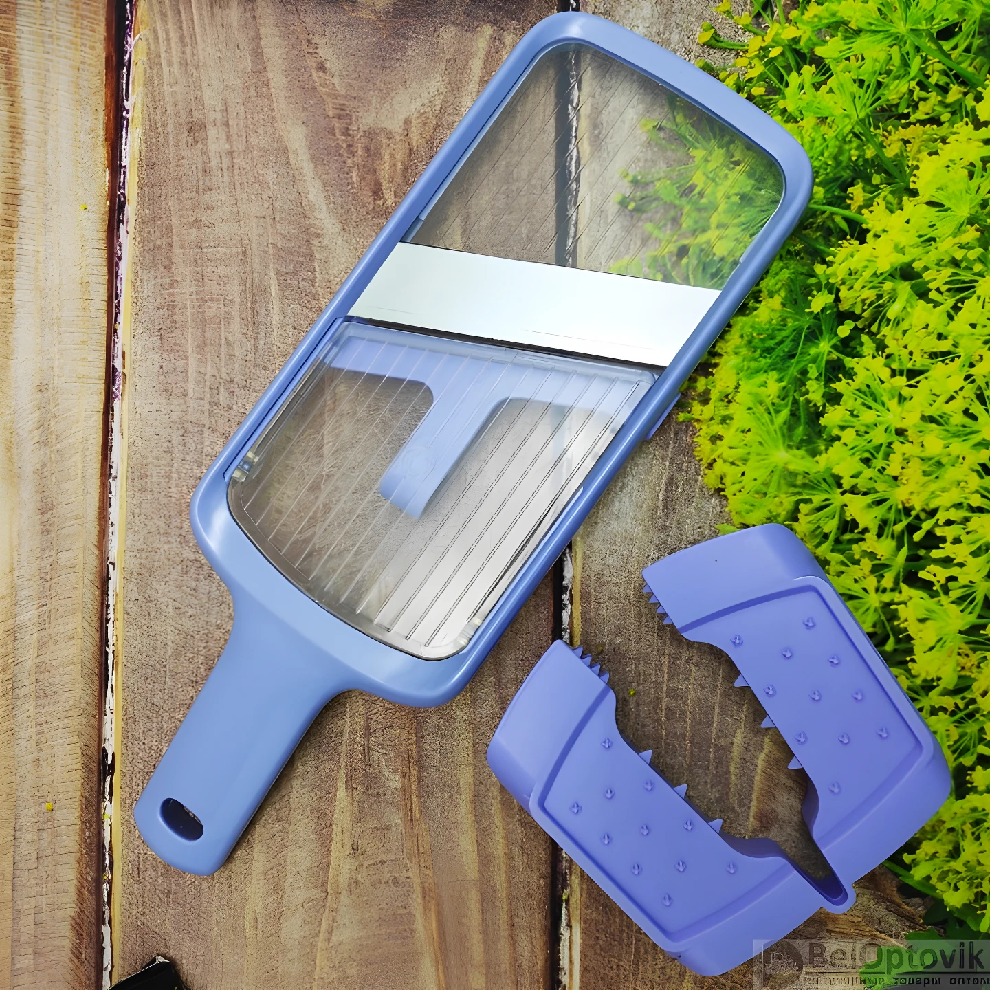 Mandolina cortadora de vegetales azul claro con cuchilla metálica y protector de agarre lila en superficie de madera y follaje verde.